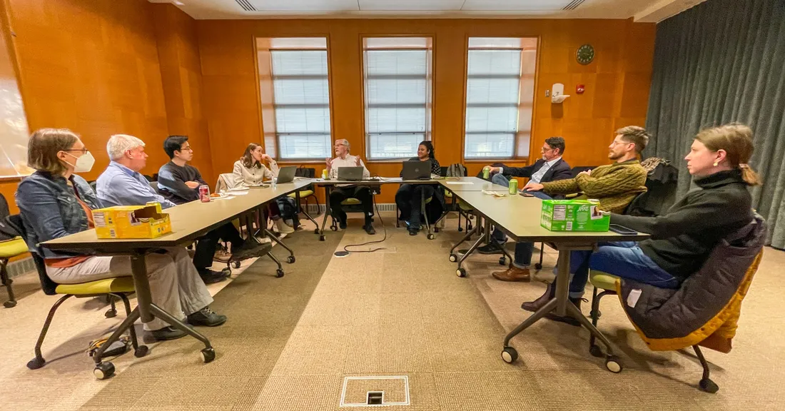 Faculty and postdocs seated around U-shaped tables in a wood-panelled meeting room as a presenter speaks.