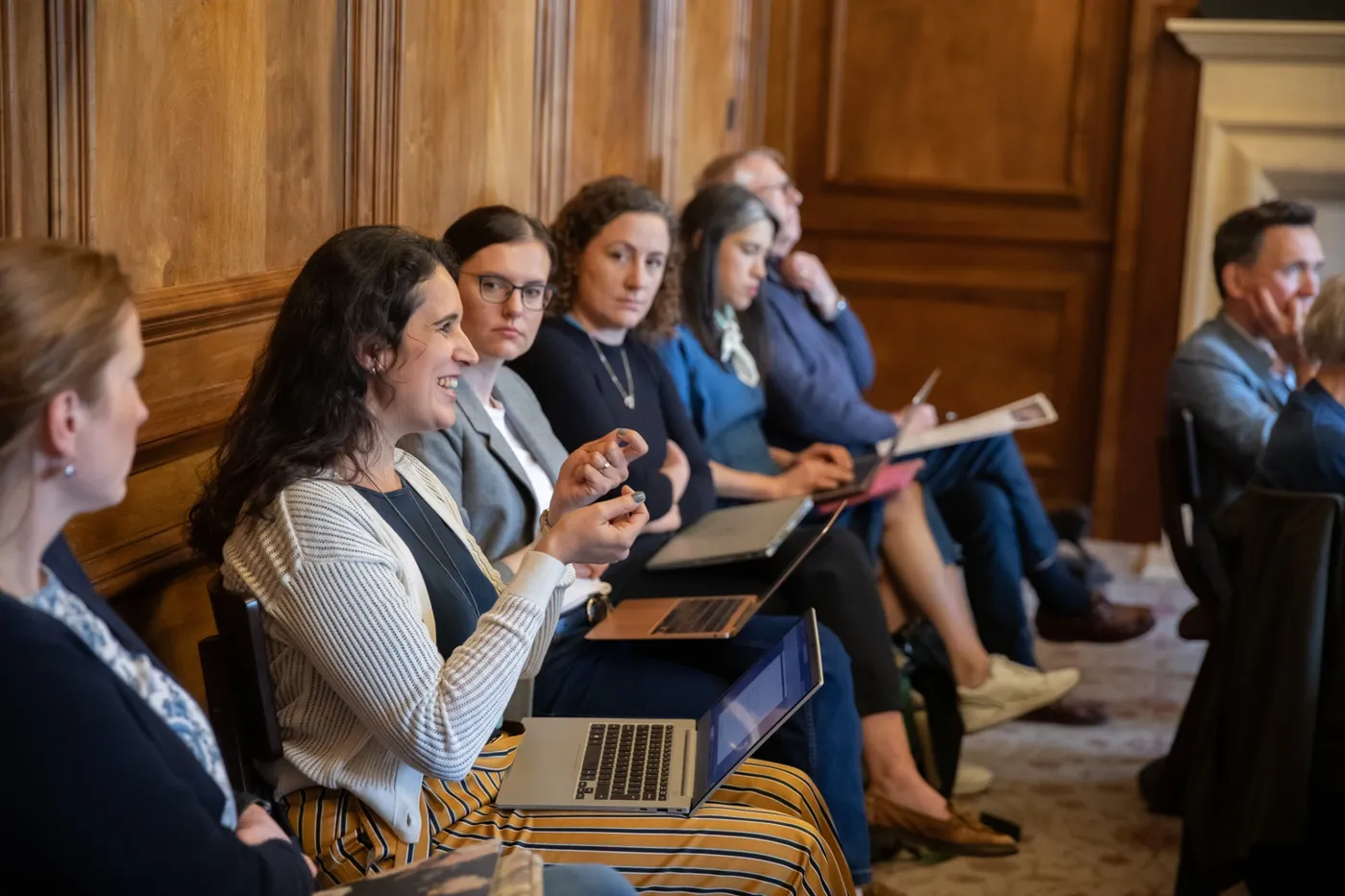 A participant speaking and gesturing during discussion while colleagues listen and take notes.