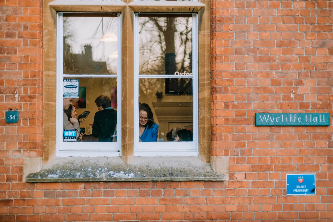 Guests gathering inside Wycliffe Hall, Oxford, seen through a window beside the hall’s nameplate.