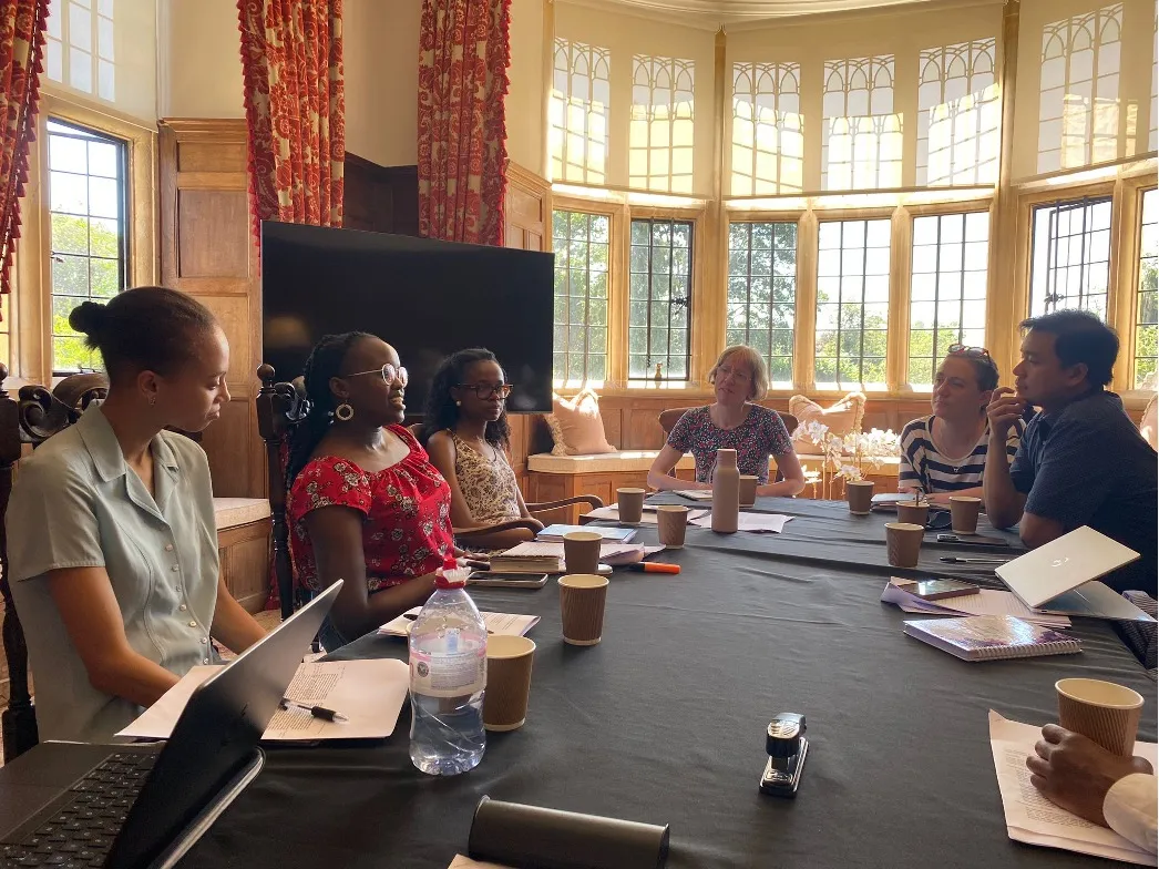 Graduate students and researchers in discussion around a table in a sunlit, wood-panelled room with tall leaded windows.