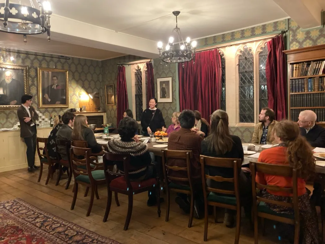 Graduate students seated around a long table in a wood-panelled Oxford dining room, listening to a speaker beneath portraits and chandeliers.
